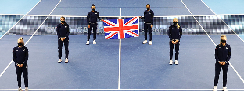 Six people standing a small distance apart on a tennis court with the back two holding a United Kingdom flag up