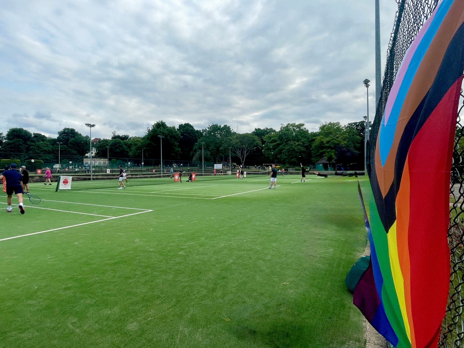 A view of multiple tennis courts with people playing on them. At the side of the image, on fencing at the back of the courts, a brighly coloured flag is attached to fencing