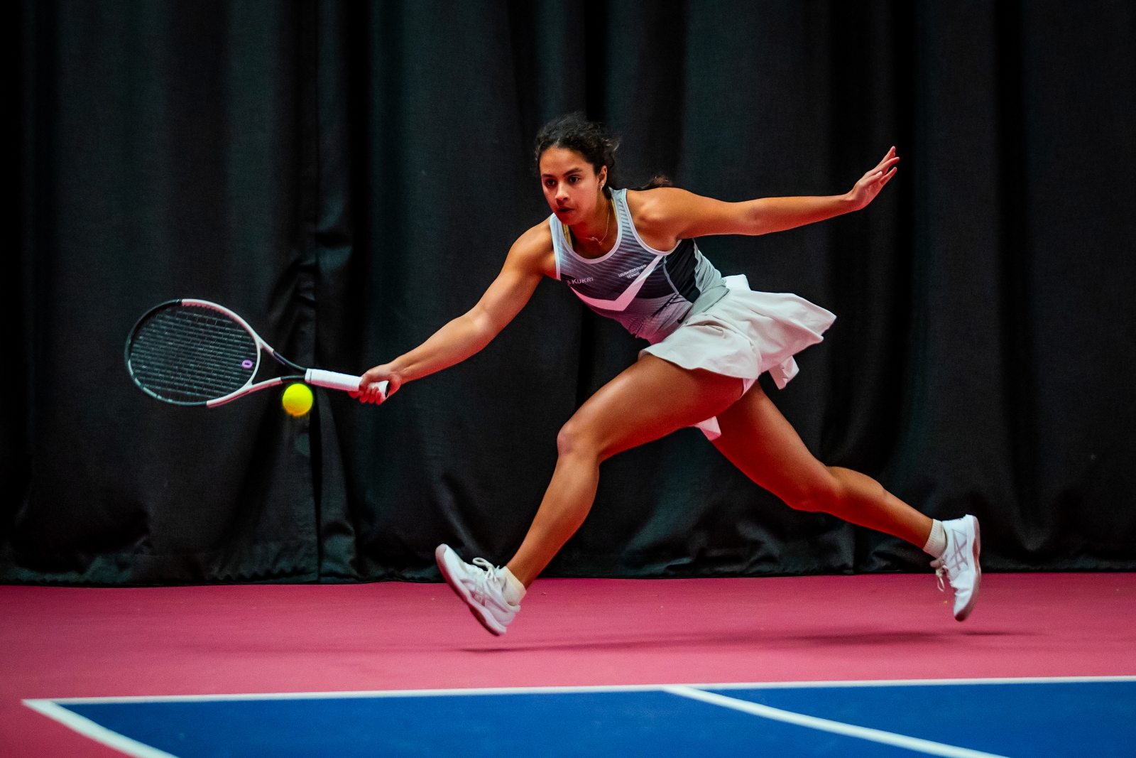 A woman runs to hit a tennis ball. She is wearing a grey vest top, a white skort and white trainers. The edge of a blue tennis court surface is visible, while she runs on a pink coloured floor outside of the court lines
