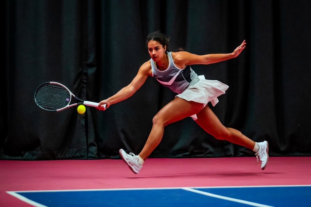 A woman runs to hit a tennis ball. She is wearing a grey vest top, a white skort and white trainers. The edge of a blue tennis court surface is visible, while she runs on a pink coloured floor outside of the court lines