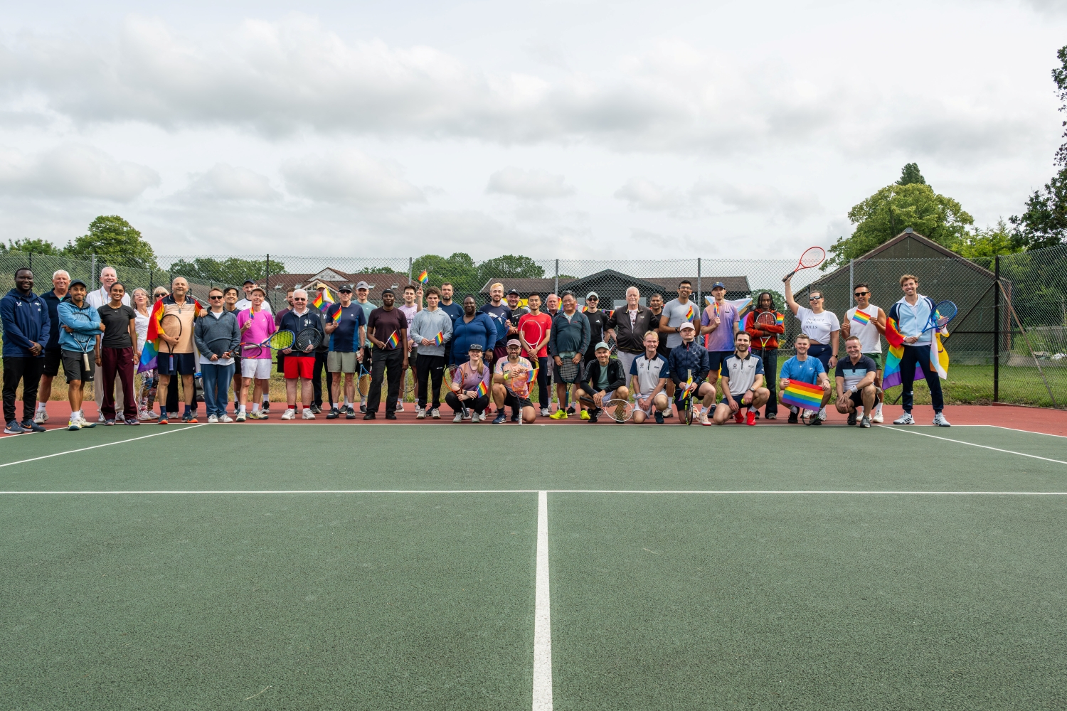 A large crowd of people stand on a tennis court, holding lots of small pride flags