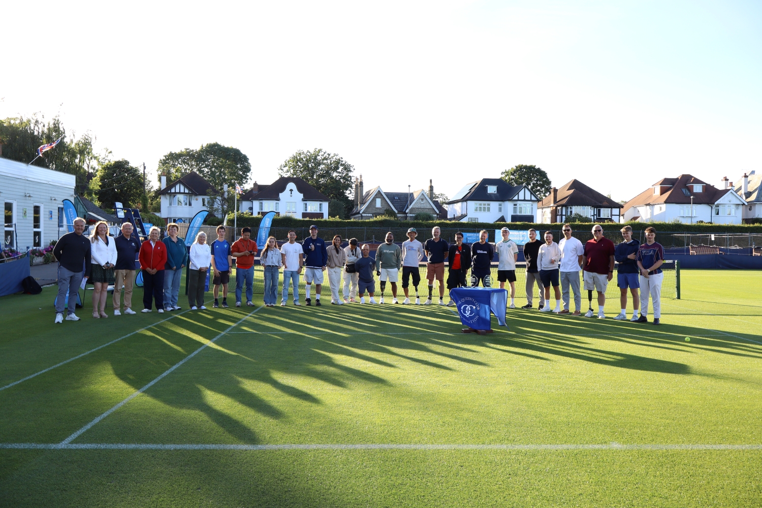 A group of people stand on a grass tennis court. Long shadows are being cast on the floor.