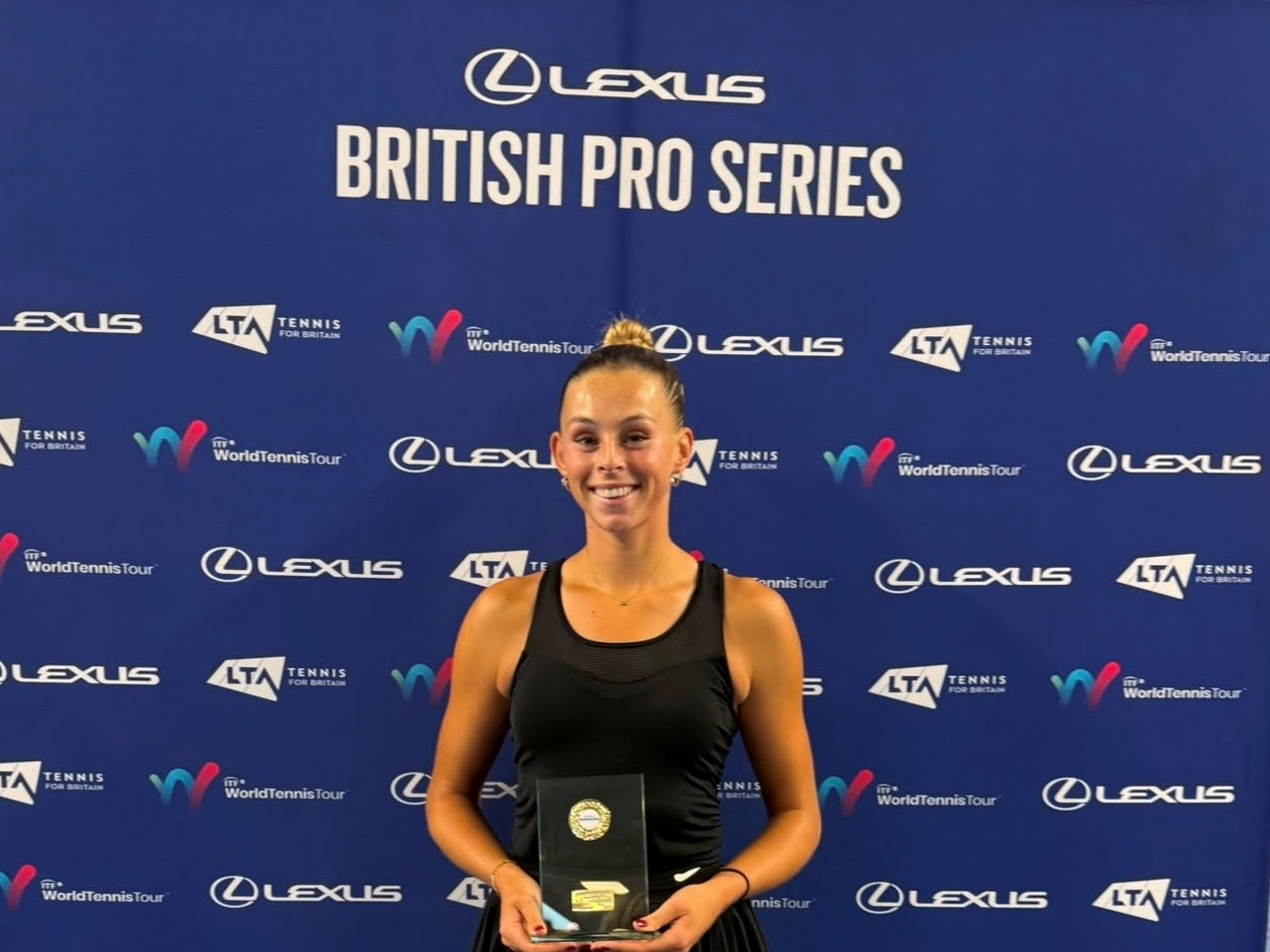 A woman wearing a black vest top satnds smiling and holding a glass trophy. Behind her is a blue backdrop featuring a variety of sponsor logos, with the words Lexus British Pro Series at the top
