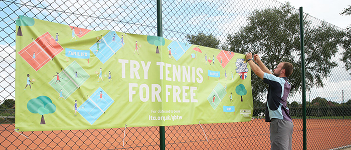 Male player attaching banner to courtside fence, that reads 'Try Tennis for Free'